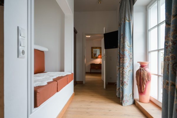 Einblick in Suite mit Durchgang, Fenster, großer Vase, Sitzgelegenheiten links und weiterem Zimmer.